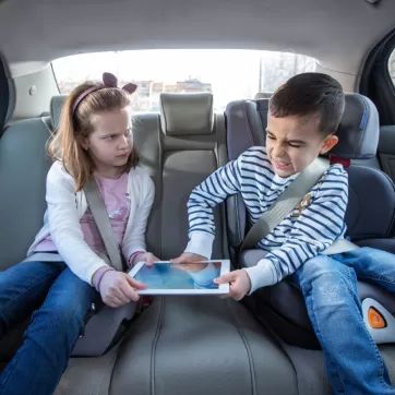 young brother and sister fighting over a tablet in the backseat of a car